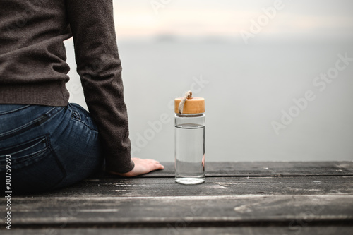 glass reusable bottle for water, stands on a wooden pier against the background of a cloudy sky next to a sitting figure of a girl
