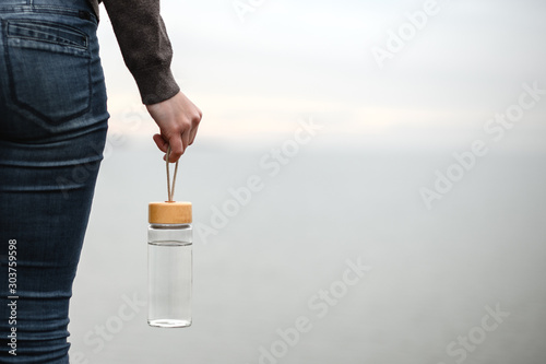 glass reusable water bottle hanging in a female hand on a background of cloudy sky