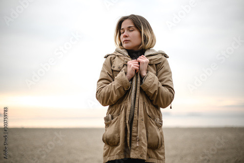 young beautiful girl with closed eyes in a warm winter jacket stands on the sand at the empty beach looks away on the background of the sea and sunset