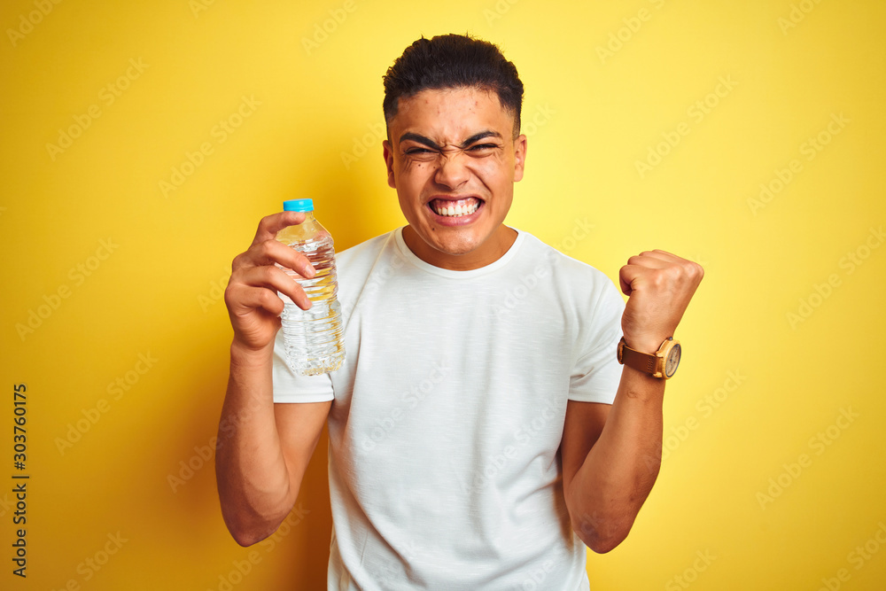 Young brazilian man holding bottle of water standing over isolated yellow background screaming proud and celebrating victory and success very excited, cheering emotion - obrazy, fototapety, plakaty 