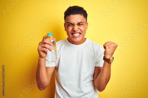 Young brazilian man holding bottle of water standing over isolated yellow background screaming proud and celebrating victory and success very excited, cheering emotion