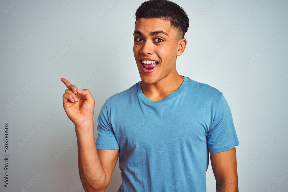 Young brazilian man wearing blue t-shirt standing over isolated white background with a big smile on face, pointing with hand and finger to the side looking at the camera. Young brazilian man wearing blue t-shirt standing over isolated white background with a big smile on face, pointing with hand and finger to the side looking at the camera.