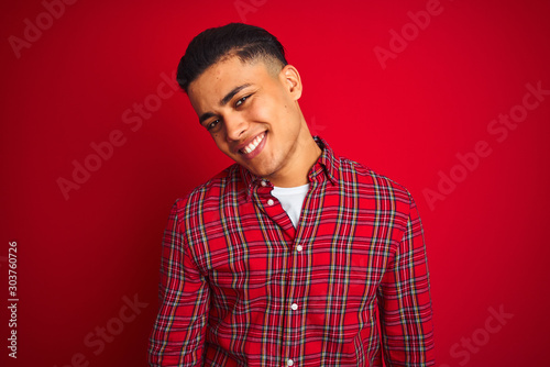 Young brazilian man wearing shirt standing over isolated red background looking away to side with smile on face, natural expression. Laughing confident.