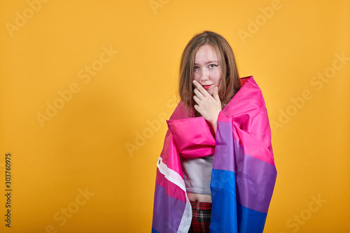 Boring young caucasian woman posing isolated on orange background in studio wearing casual shirt, keeping hand on chin, covered bisexual flag