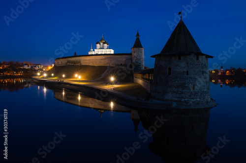 Night panorama of the city. Lanterns illuminate the walls and towers of the medieval fortress and are reflected in the water of the river. Pskov, Russia.