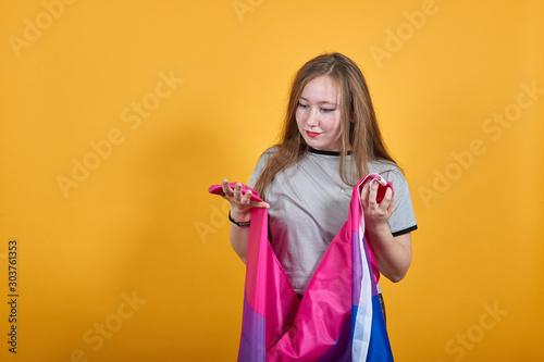 Smile cheerful young woman in casual shirt  looking down  spreading hands covered bisexual flag isolated on orange background in studio. People sincere emotions  lifestyle concept.