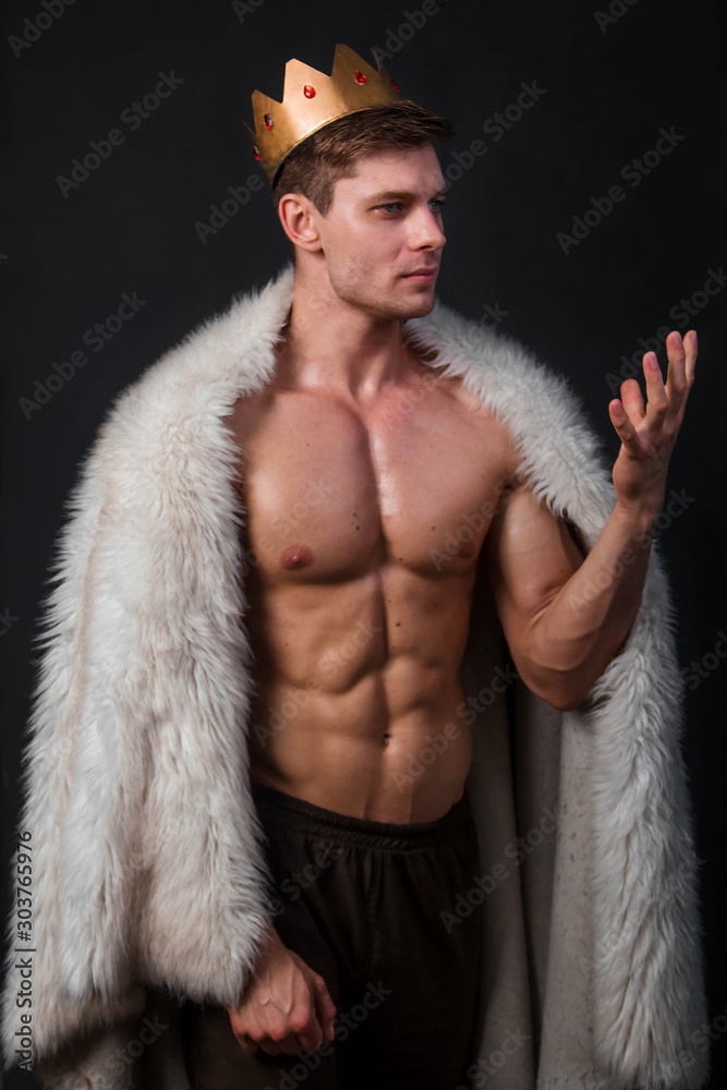 Portrait of a young bodybuilder with a crown on his head and a fur gown on his shoulders. Studio photo on a black background. Portrait of a young bodybuilder with a crown on his head and a fur gown on his shoulders. Studio photo on a black background.