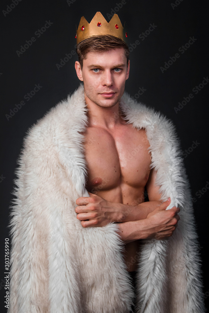 Portrait of a young bodybuilder with a crown on his head and a fur gown on his shoulders. Studio photo on a black background.