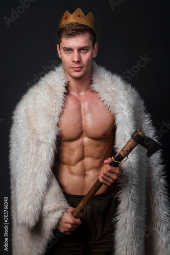 Portrait of a young bodybuilder with a crown on his head and a fur gown on his shoulders and an ax in his hands. Studio photo on a black background.