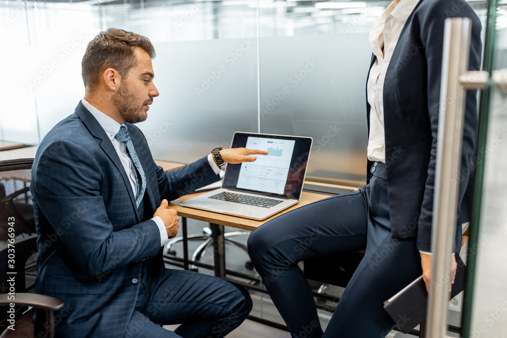 Group of strictly dressed business people discussing financial graph presented on the laptop, meeting in the modern office with glass partitions Group of strictly dressed business people discussing financial graph presented on the laptop, meeting in the modern office with glass partitions