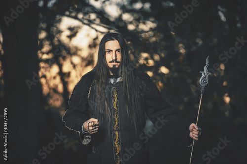 A man with long hair and beard in a black cloak in the image of a sorcerer warlock on Halloween. Dark magician the man in the background of the forest. A wizard in a dark forest with a spear