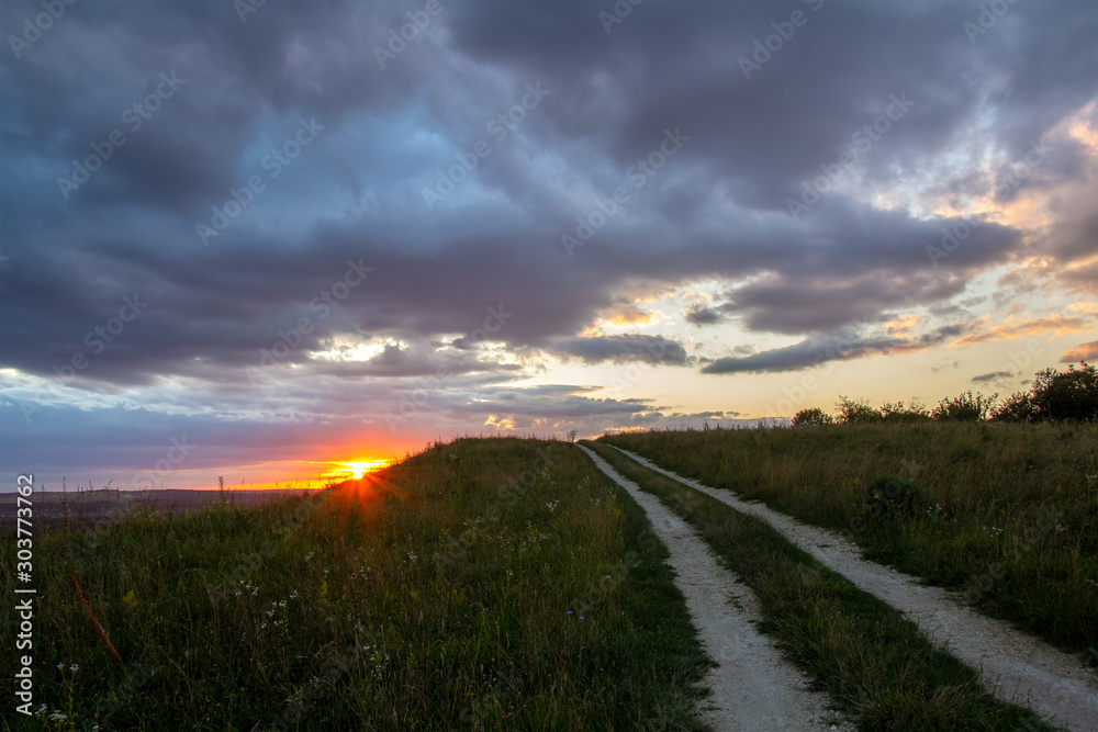 Dirt road among dark fields at sunset with dramatic cloudscape.