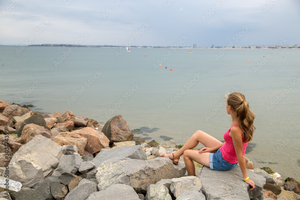 Young woman in summer clothes sitting on big boulders on sea shore looking on horizon.