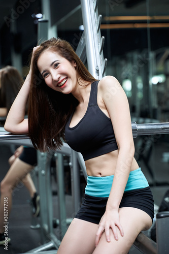 A happy young beautiful woman is sitting on a barbell rack and having a good chat.