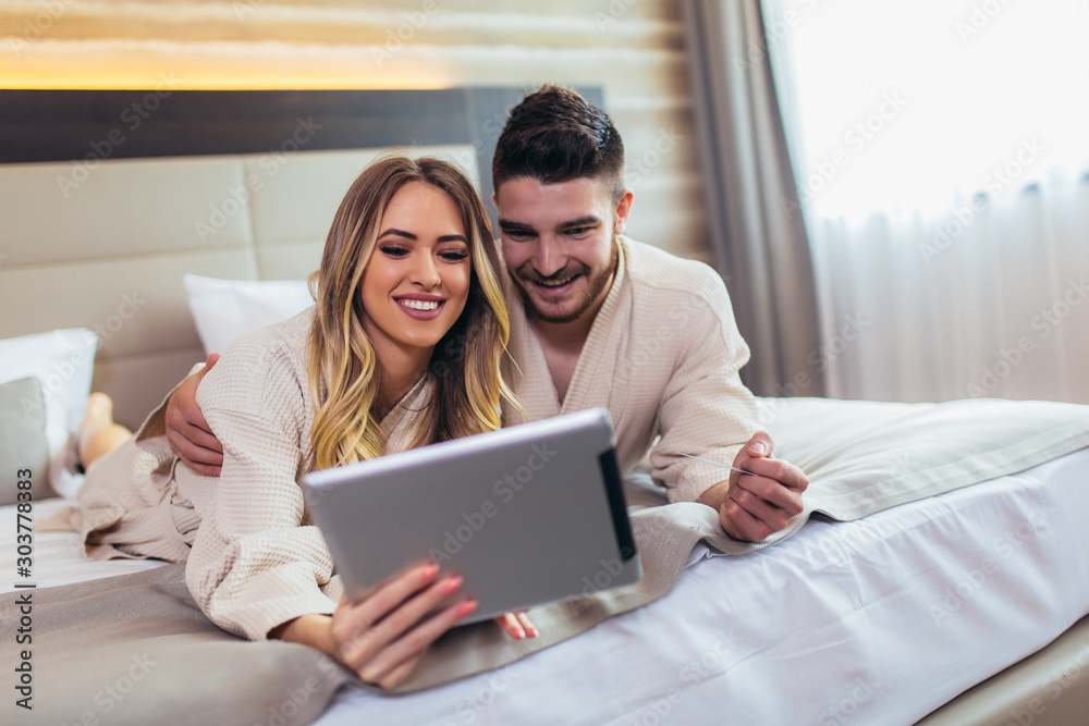 Happy couple using digital tablet while lying together in bed in hotel room. Happy couple using digital tablet while lying together in bed in hotel room.