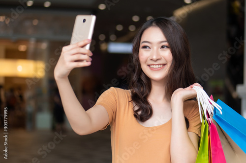 happy smiling woman shopping using smartphone while holding shopping bag in shopping mall; concept of smartphone upload, social network influencer, grand sale discount, good deal for woman shoppers