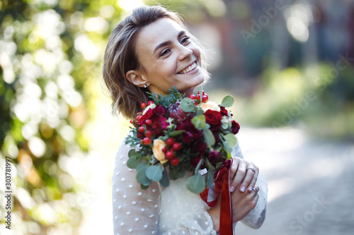 Beautiful bride with a wedding bouquet for a walk in the park. Young bride in the fashionable white dress is incredibly happy. Elegant woman with professional make up and hair style. Wedding day. 