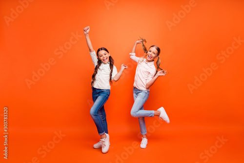 Full body photo of funky crazy two girlfriends have fun on fall weekends raise hands fists hold arms pigtails wear white shirt sneakers isolated orange color background