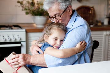 Grandmother congratulates child with gift box. Kid and senior woman are hugging in cozy home kitchen. Family enjoying kindness, tenderness. Lifestyle moment. Happy holidays Christmas, Birthday