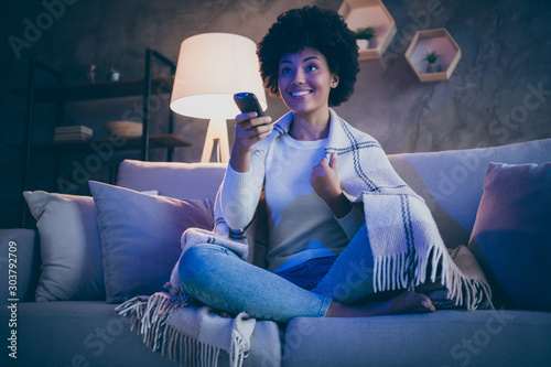 Cheerful afro american girl having free time watch tv film hold remote control switch channels sit divan crossed legs cover checkered blanket in evening room house indoors