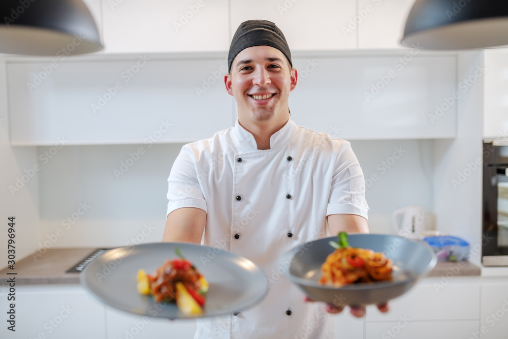 Smiling caucasian creative chef in white uniform holding plates with tagliatelle and salmon with orange while standing in kitchen. Smiling caucasian creative chef in white uniform holding plates with tagliatelle and salmon with orange while standing in kitchen.