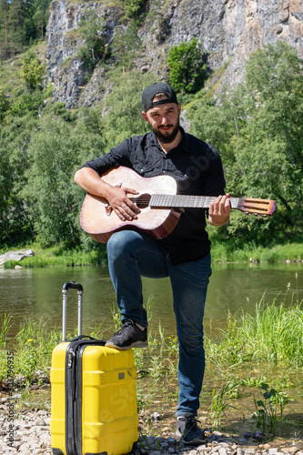 Hitchhiking guy. Concept of travel, holidays, vacation. Man with guitar on background of mountains, forests and lakes.