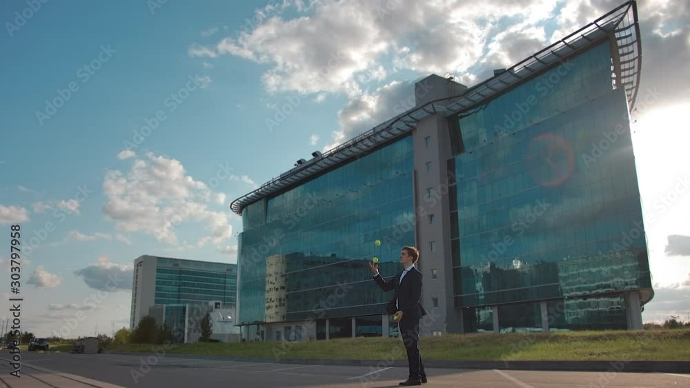 Calm and confident juggling businessman. Well dressed person in formal suit standing outdoors. Stable, reliable and control. Overall plan with office building on the background Calm and confident juggling businessman. Well dressed person in formal suit standing outdoors. Stable, reliable and control. Overall plan with office building on the background