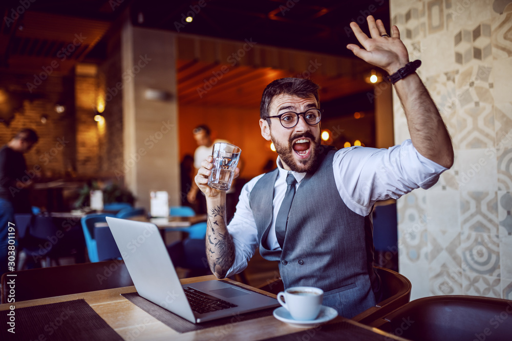 Attractive caucasian bearded businessman in suit and with eyeglasses holding glass of water and waving while sitting in cafe. On table are laptop and cup of coffee. - obrazy, fototapety, plakaty 