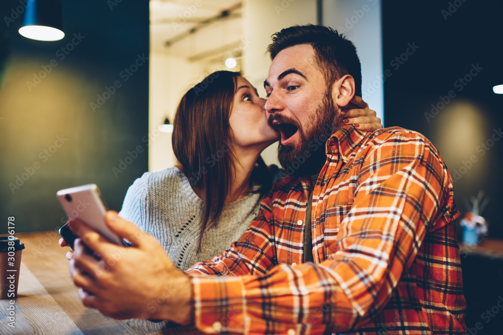 Happy young woman kisses her bearded boyfriend who won prize in internet lottery.Amazed coung couple in love rejoices in victory in online contest on smartphone device sitting at home interior - obrazy, fototapety, plakaty 