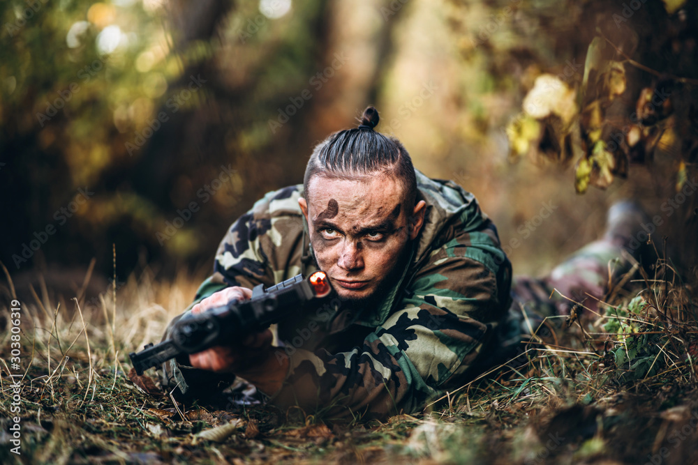 Portrait of a camouflage soldier with rifle and painted face lying in the grass aiming at the rifle. Playing airsoft outdoors in the forest