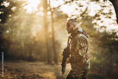 A camouflage soldier playing airsoft outdoors in the forest, sunset time, aiming at the rifle