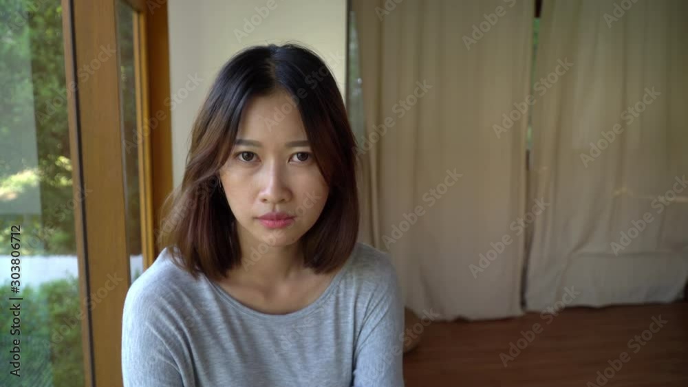 4K Depressed young beautiful asian woman face looking at camera. Loneliness woman with sadness eye sitting alone in the room near the window at home. Portrait woman face and mental health care concept 4K Depressed young beautiful asian woman face looking at camera. Loneliness woman with sadness eye sitting alone in the room near the window at home. Portrait woman face and mental health care concept