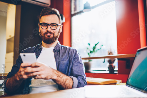 Portrait of handsome bearded male blogger looking at camera while spending leisure time in cafe interior with good internet connection, young hipster guy in eyeglasses holding smartphone gadget