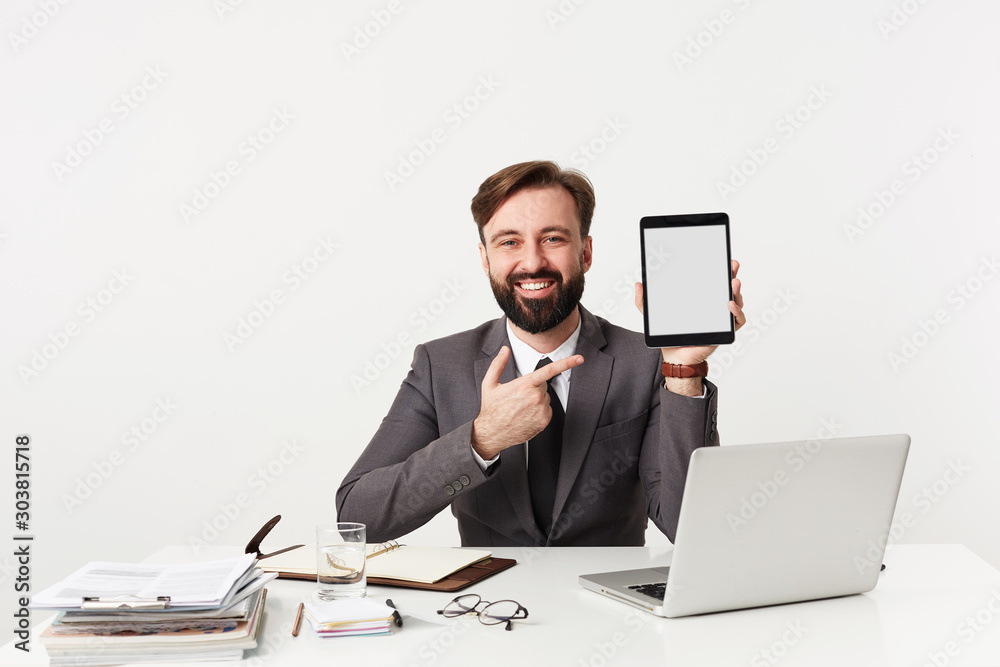 Cheerful handsome young brunette man with beard wearing formal clothes over white background while sitting at table with tablet pc in raised hand, looking happily to camera with wide smile Cheerful handsome young brunette man with beard wearing formal clothes over white background while sitting at table with tablet pc in raised hand, looking happily to camera with wide smile