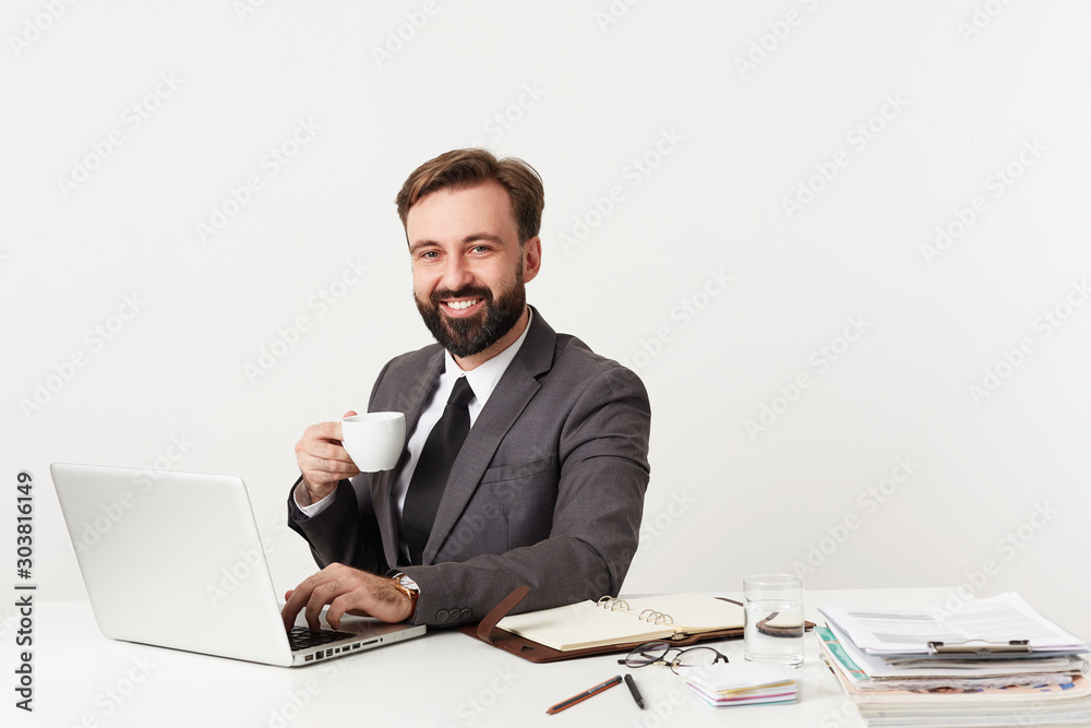 Happy attractive bearded businessman with short brown hair holding cup of tea and looking joyfully at camera, working with laptop and typing letter to his partners, isolated over white background Happy attractive bearded businessman with short brown hair holding cup of tea and looking joyfully at camera, working with laptop and typing letter to his partners, isolated over white background