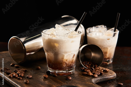 white russian cocktail in glasses with straws on wooden board with coffee grains and shaker isolated on black photo