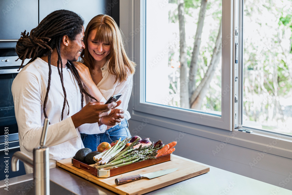 Girl leaning on the shoulder of a boy of different ethnicity in the kitchen looking at the mobile and smiling next to a window