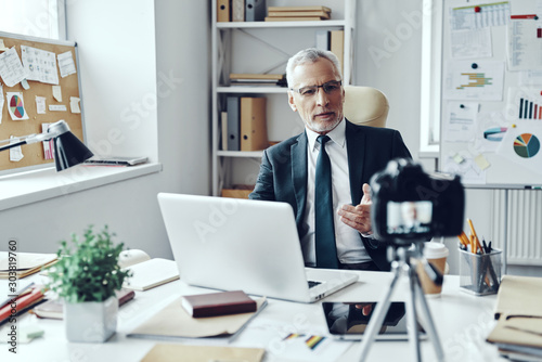 Senior man in elegant shirt and tie working using laptop while making social media video