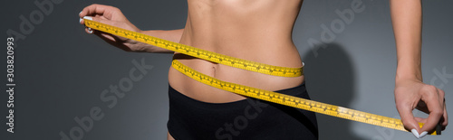 cropped view of slim woman in underwear holding measuring tape on waist on dark background, panoramic shot photo