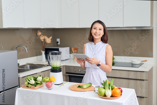 Asian woman using tablet to find recipe making smoothie with fruit and vegetable in kitchen.