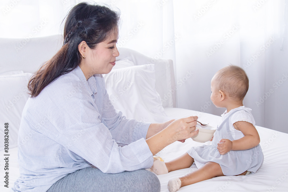 Beautiful Asian woman mother feeding fruits nutrition to her toddler girl baby by spoon, adorable baby girl eating fruits in bowl, happy motherhood relationship taking care between mother and daughter Beautiful Asian woman mother feeding fruits nutrition to her toddler girl baby by spoon, adorable baby girl eating fruits in bowl, happy motherhood relationship taking care between mother and daughter