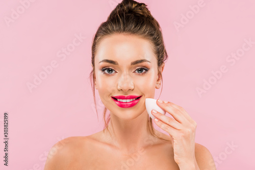 smiling naked beautiful woman with pink lips holding makeup sponge isolated on pink