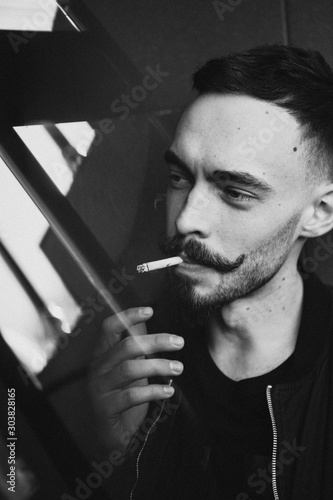 a young brutal bearded man with a stylish mustache lights a cigarette and smokes. Black and white photo
