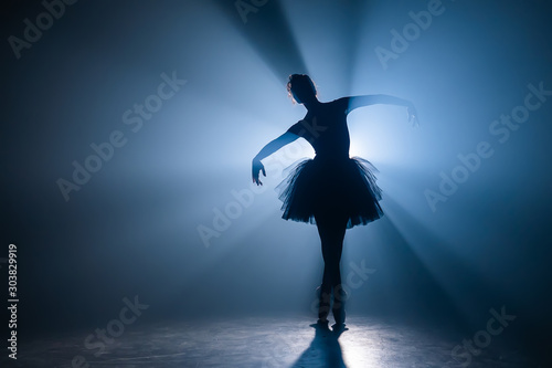 Ballerina in black tutu dress dancing on stage with magic blue light and smoke. Silhouette of young attractive dancer in ballet shoes pointe performing in dark. Copy space.