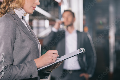selective focus of businesswoman writing on clipboard near businessman talking on smartphone in warehouse