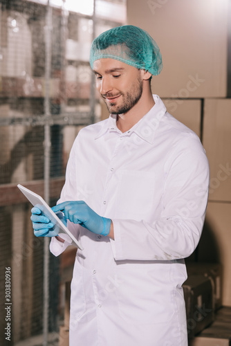Φωτογραφία smiling storekeeper in white coat and hairnet using digital tablet in warehouse