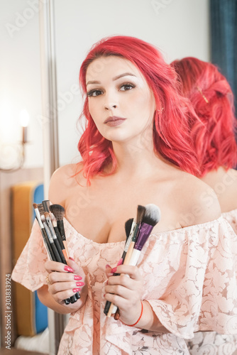Portrait of the beautiful woman with make-up brushes near attractive face. Adult girl posing over white background in her room alone