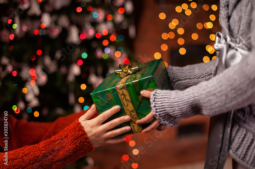 Hands give and receive beautiful wrapped christmas present on colorful blurry lights background