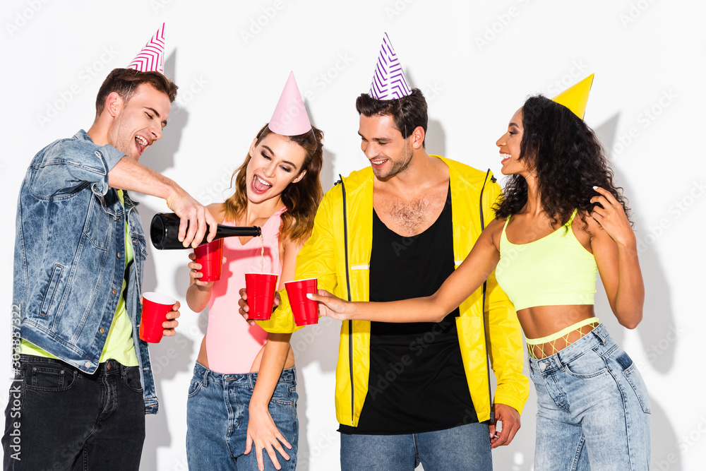 Φωτογραφία happy man pouring champagne in plastic cups near multicultural friends in party