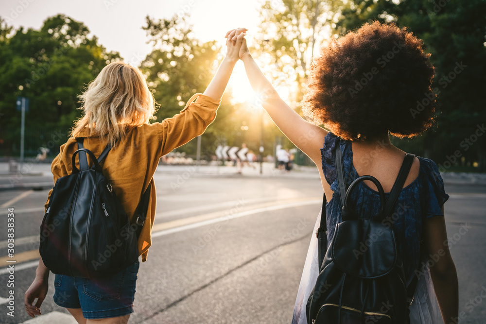 Couple of young women from the back, holding hands with arms raised and they walk in the street at sunset - Two millennials are happy - obrazy, fototapety, plakaty 